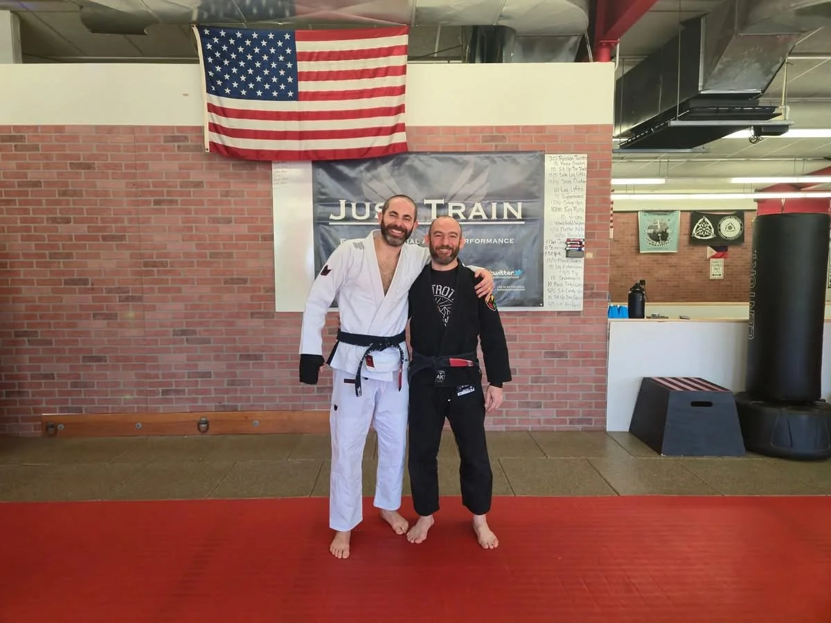 Two men in jiu-jitsu gis, one in a white gi and the other in a black gi, standing on a red mat inside a gym, smiling with their arms around each other. An American flag hangs on the wall behind them along with a banner that says "Just Train." There are also some posters, a whiteboard with writing, and gym equipment visible in the background.