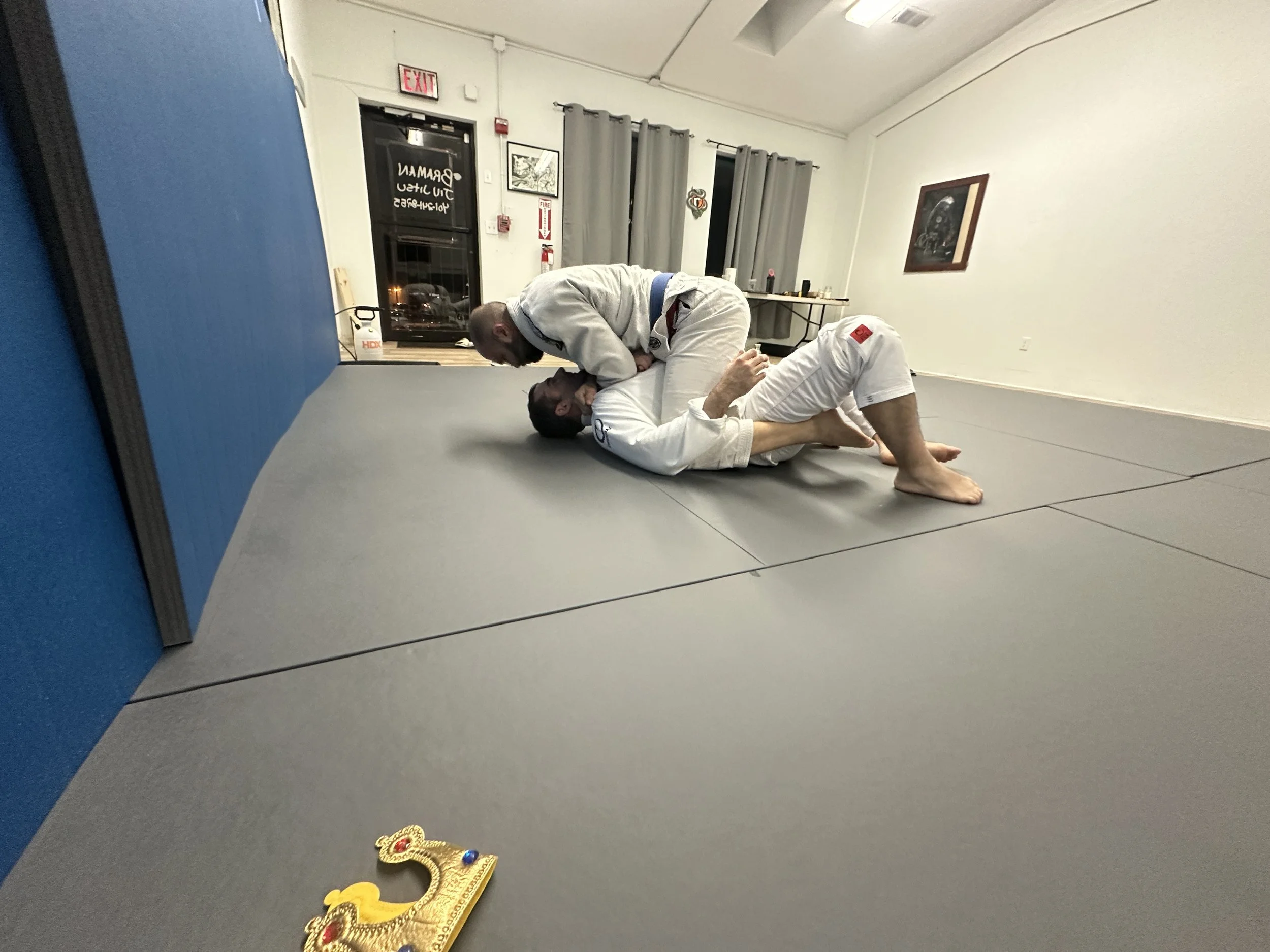 Two men practicing Brazilian Jiu-Jitsu on a gray mat in a martial arts gym, with one man in a white gi on top and the other in a white gi on bottom, engaged in a grappling hold.
