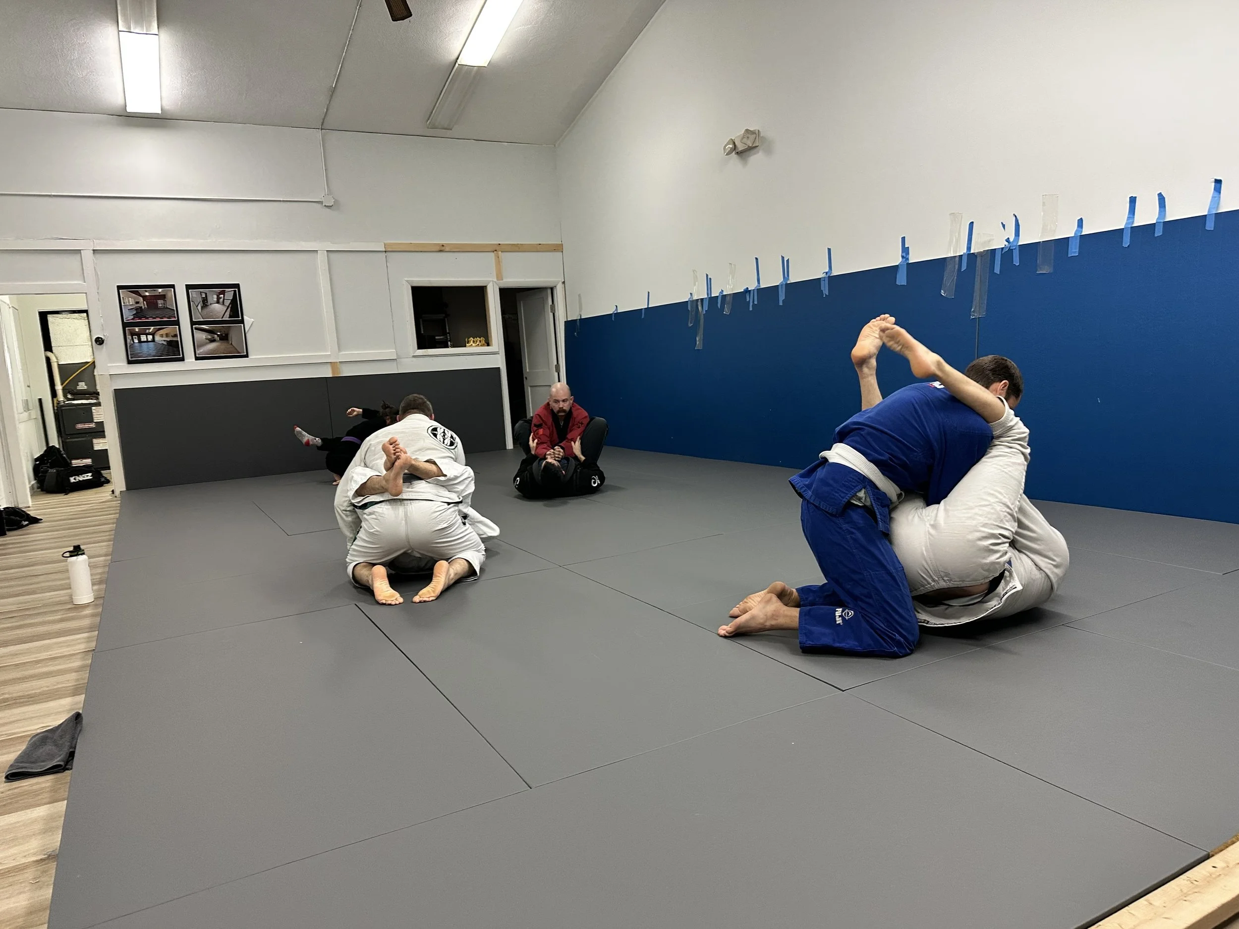 Two pairs of people practicing Brazilian Jiu-Jitsu on gray mats in a training room, with a referee or instructor observing, and a white and blue wall in the background with taped line markings.