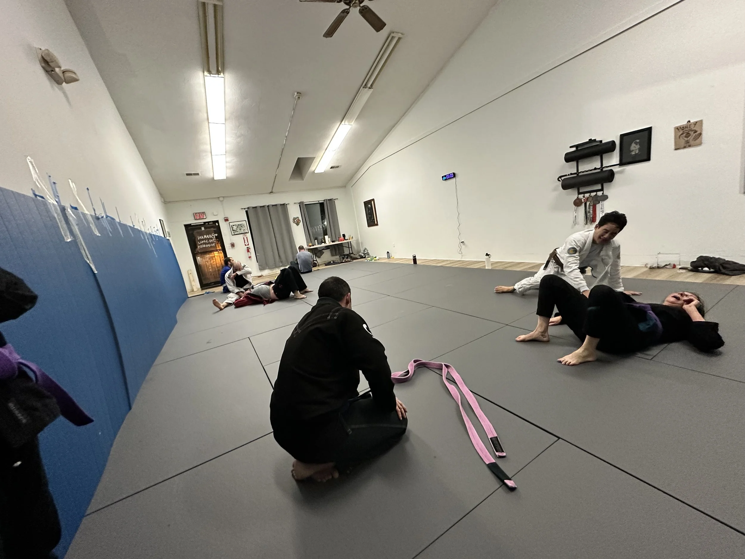 Martial arts students practicing on mats in a gym, with one instructor observing. The room has white walls, gray mats, and dark-colored equipment on the walls.