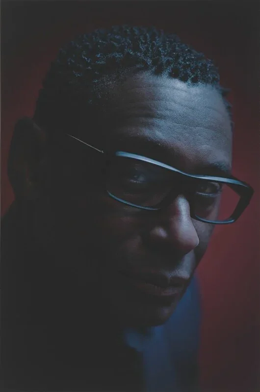 Close-up of a man with short, curly hair wearing glasses, looking over his shoulder against a dark red background.