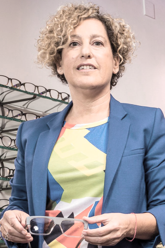 A woman with curly blonde hair holding prescription eyeglasses in an optical store, standing in front of a display of eyeglasses.