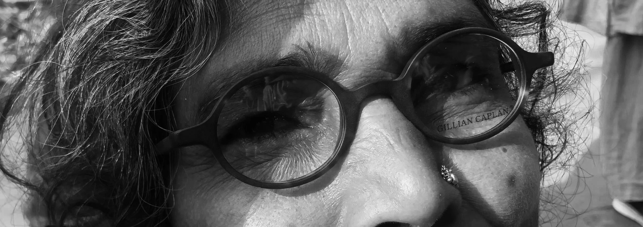 Close-up of a person's face with curly hair wearing round glasses with the name 'Gillian Caplan' on the lens. Black and white photo.