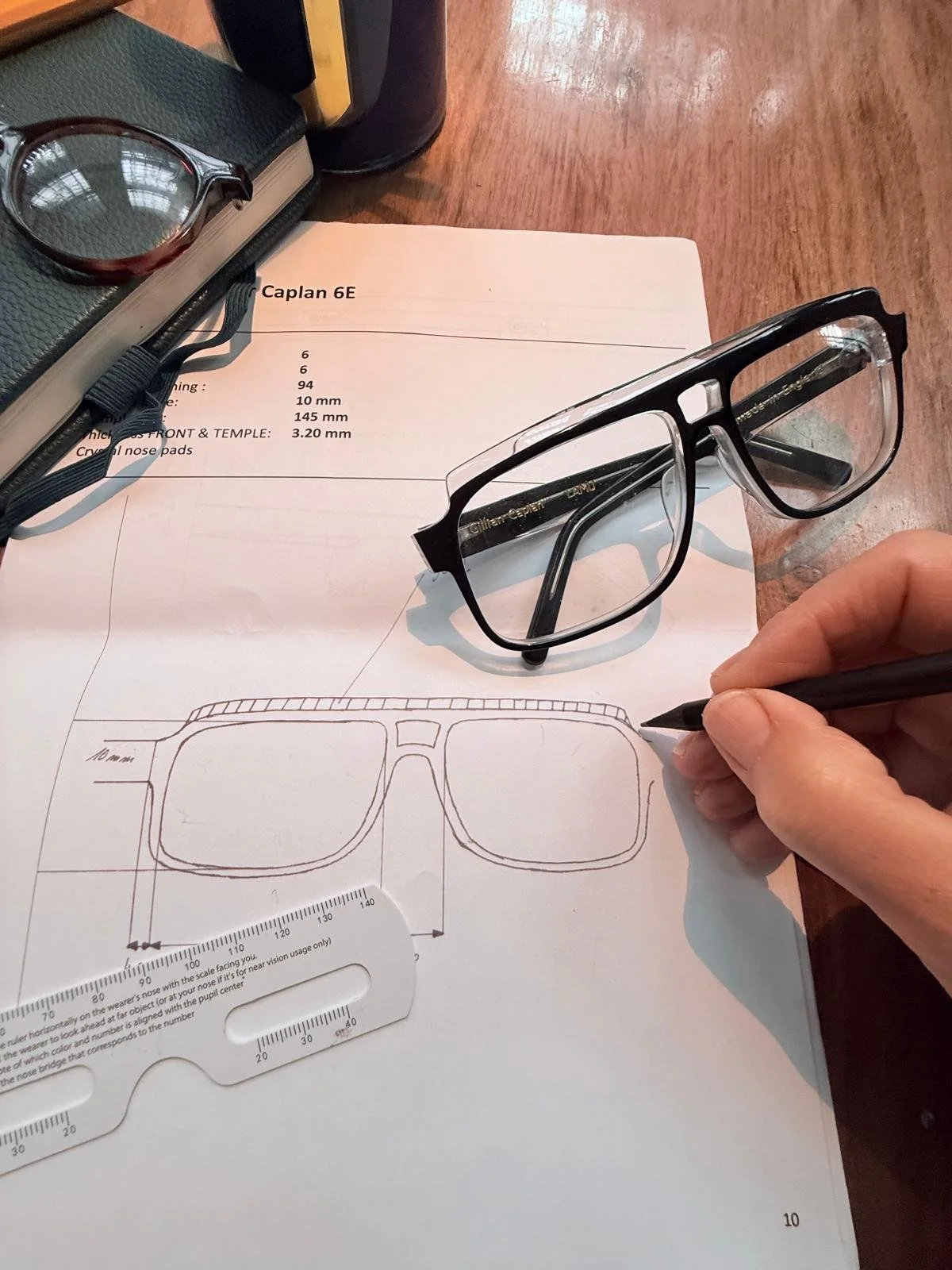 A person is sketching a drawing of eyeglasses on a piece of paper. A real pair of eyeglasses is resting on the paper, and a ruler is nearby. There are other glasses and books on the wooden table.