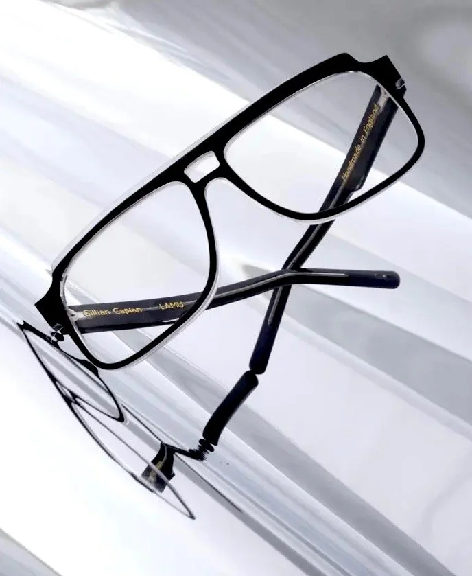 A pair of black eyeglasses with a gold inscription on the inside of the arm resting on a reflective surface.