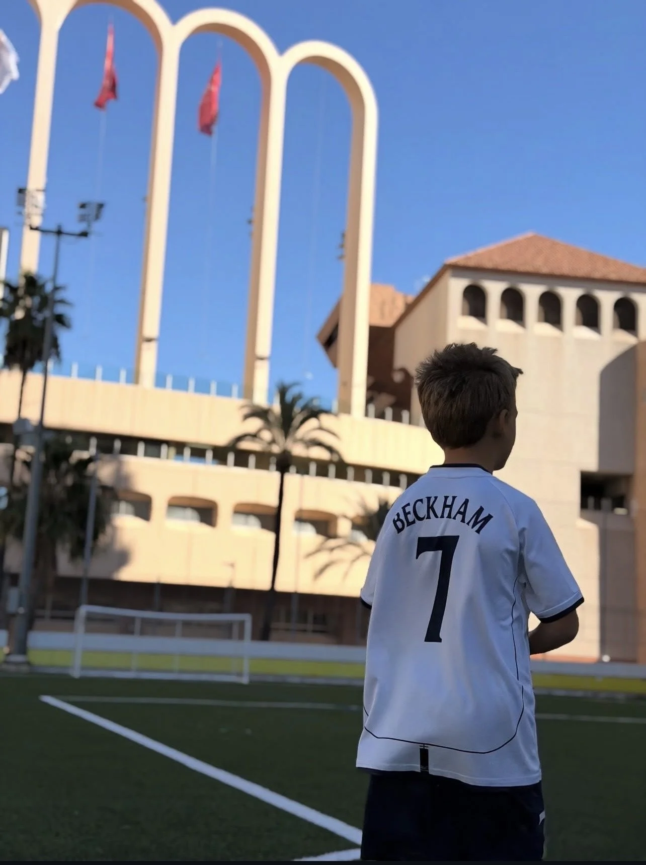 A young boy stands on a soccer field wearing a white Beckham jersey with the number 7, facing away from the camera. In the background, there are palm trees, a multi-story building with arched windows, and a large structure with tall arches and flags,