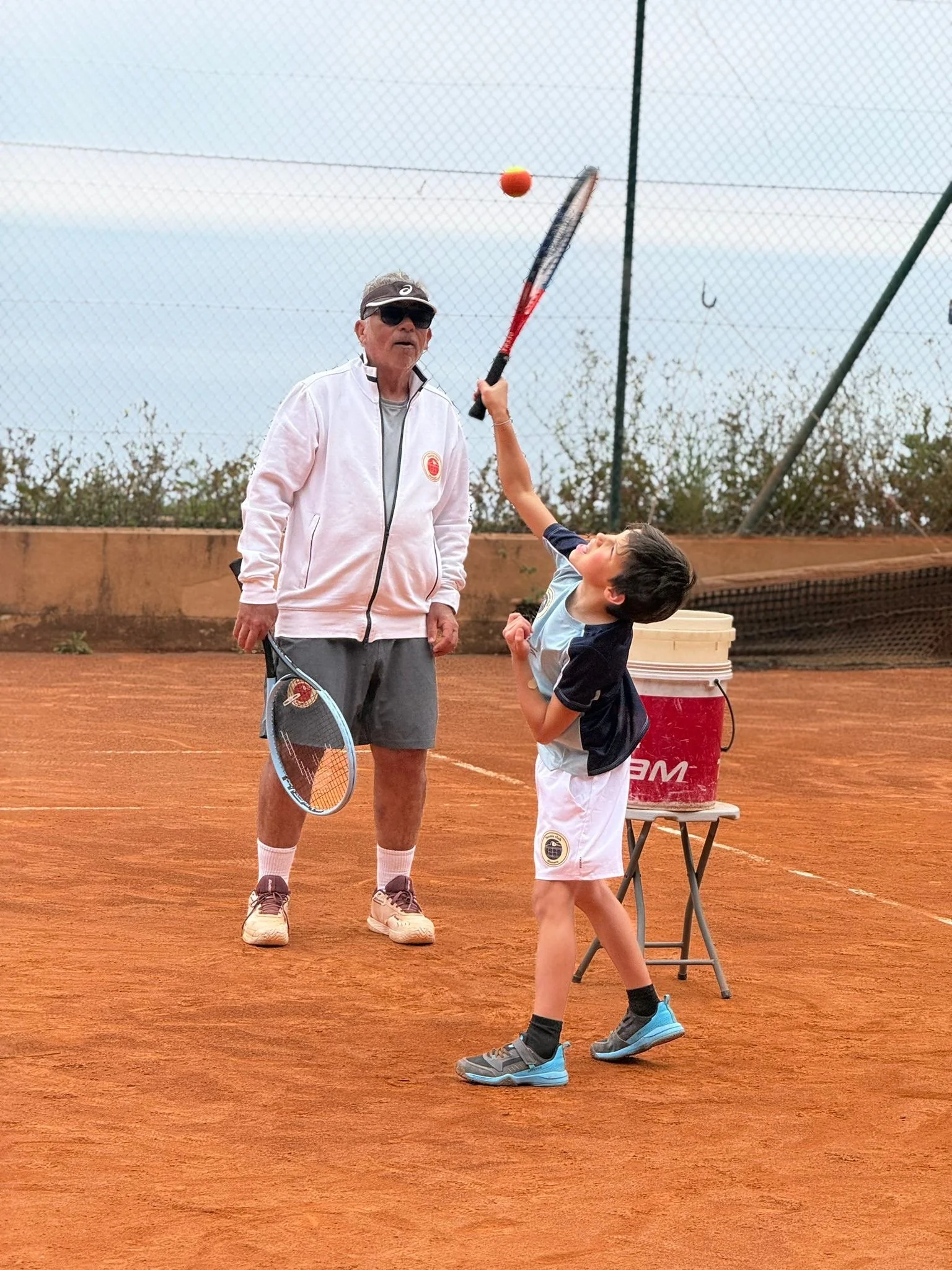 An elderly man and a young boy playing tennis on a clay court. The man is wearing sunglasses, a white jacket, gray shorts, and shoes, while the boy is wearing a black and white shirt, white shorts, and gray sneakers. There is a table with a red cooler in the background, and the court is enclosed with a chain-link fence.