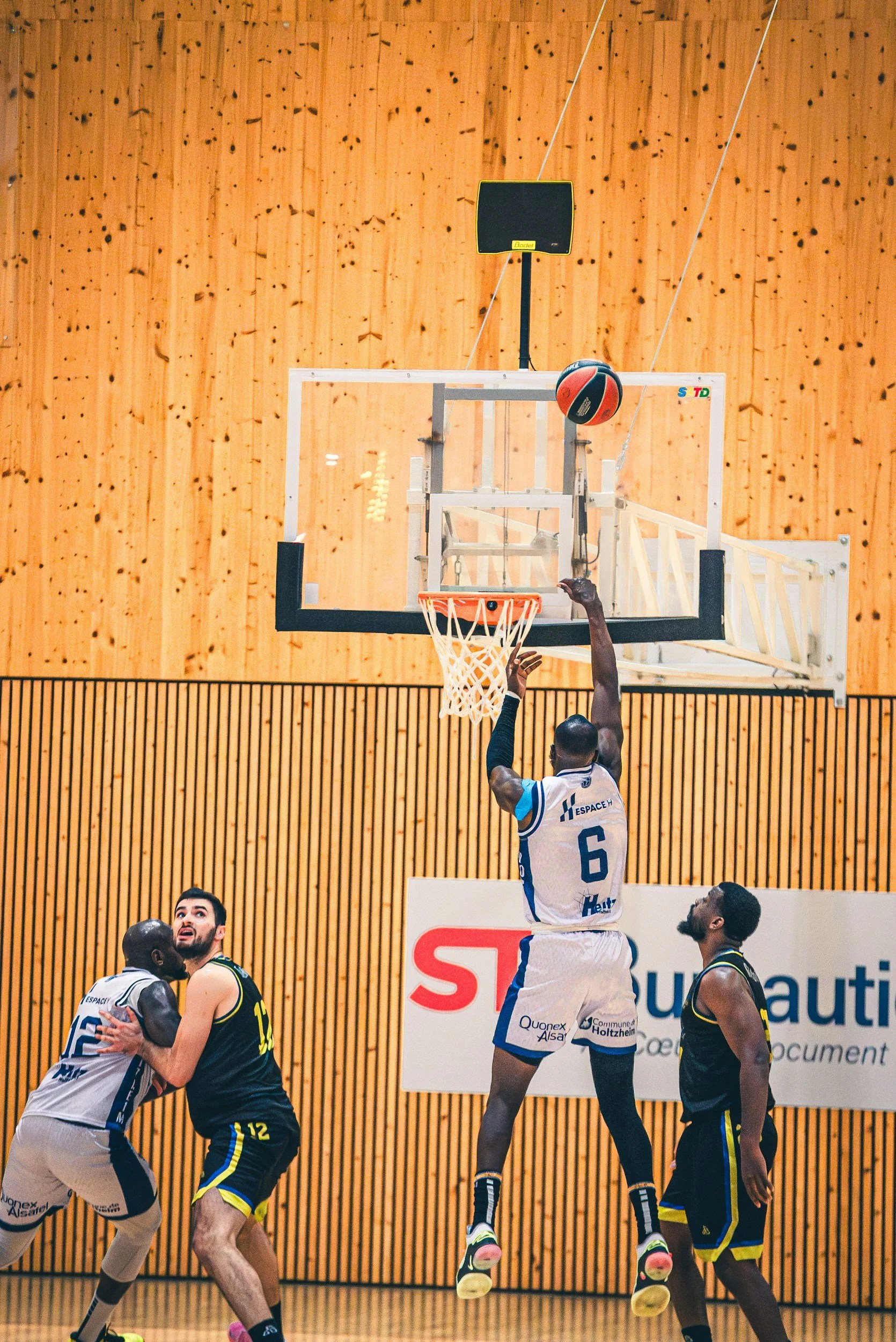 A basketball game in action with players jumping for the ball near the hoop, in a gym with wooden wall panels.