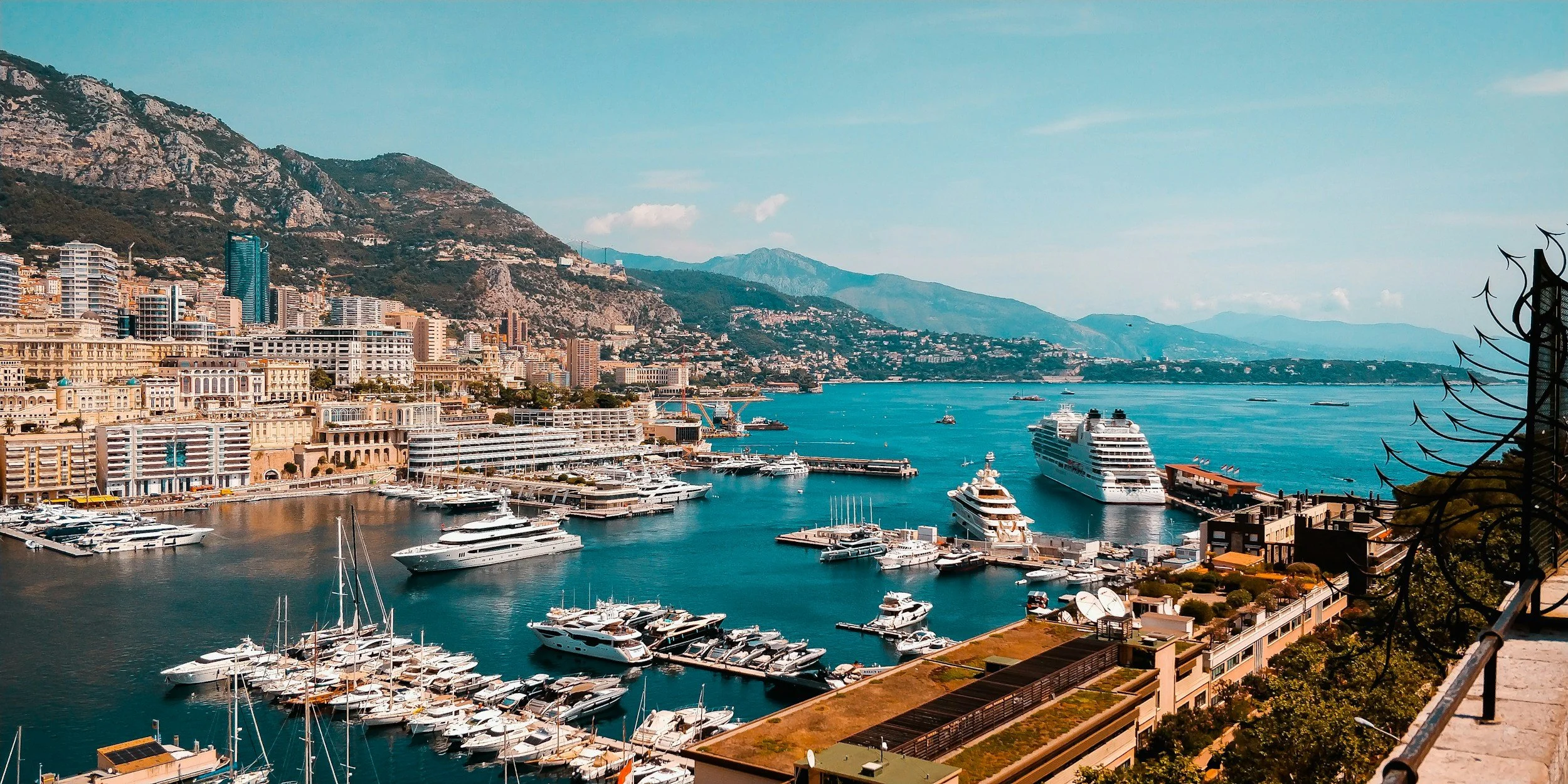 A panoramic view of a busy marina with yachts and boats docked along the waterfront, surrounded by high-rise buildings, mountains in the background, and a clear blue sky.