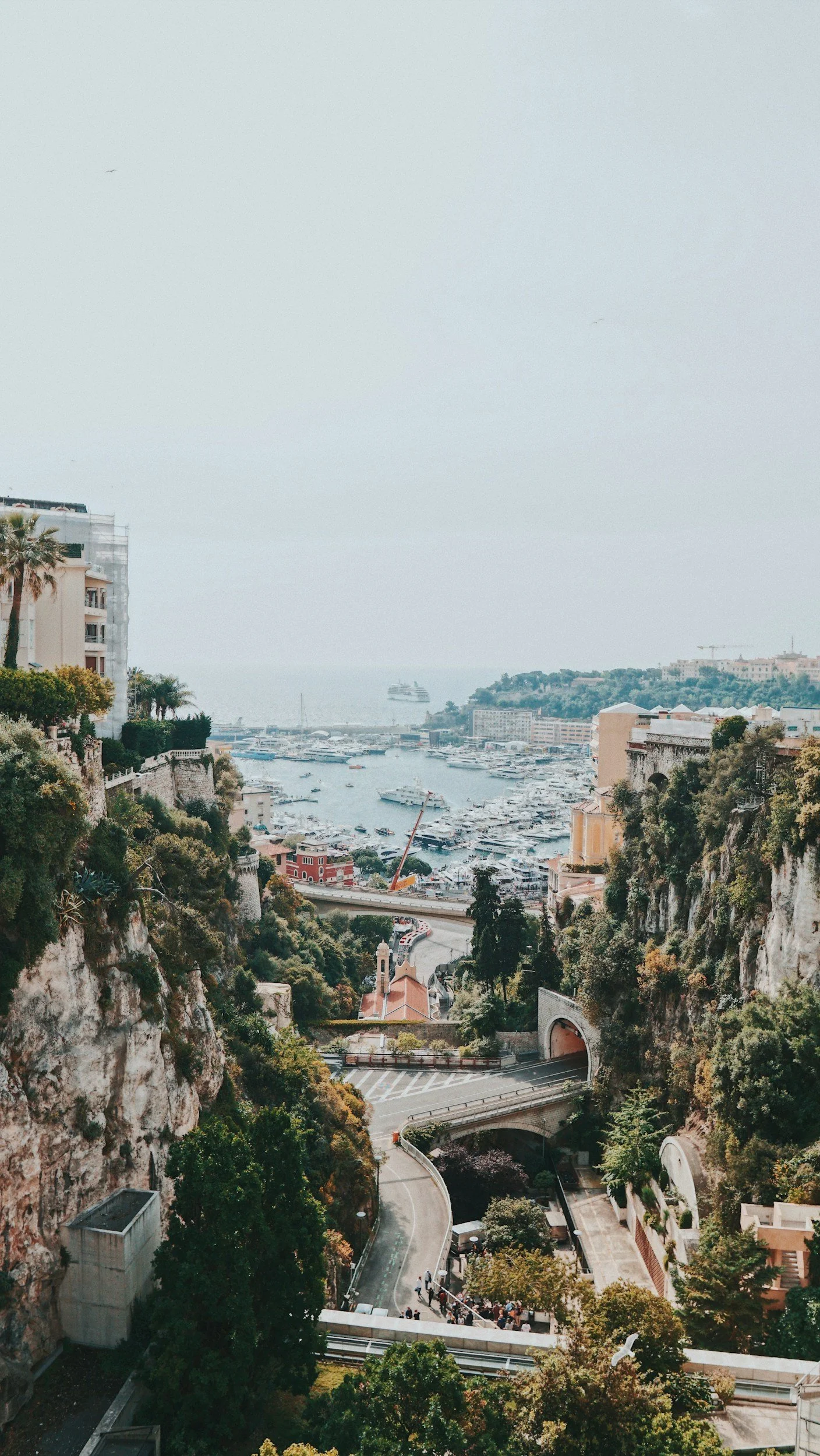 Scenic view of a coastal city with a harbor filled with boats, surrounded by hills with lush greenery and buildings, a cruise ship in the water, and a winding road with people walking.