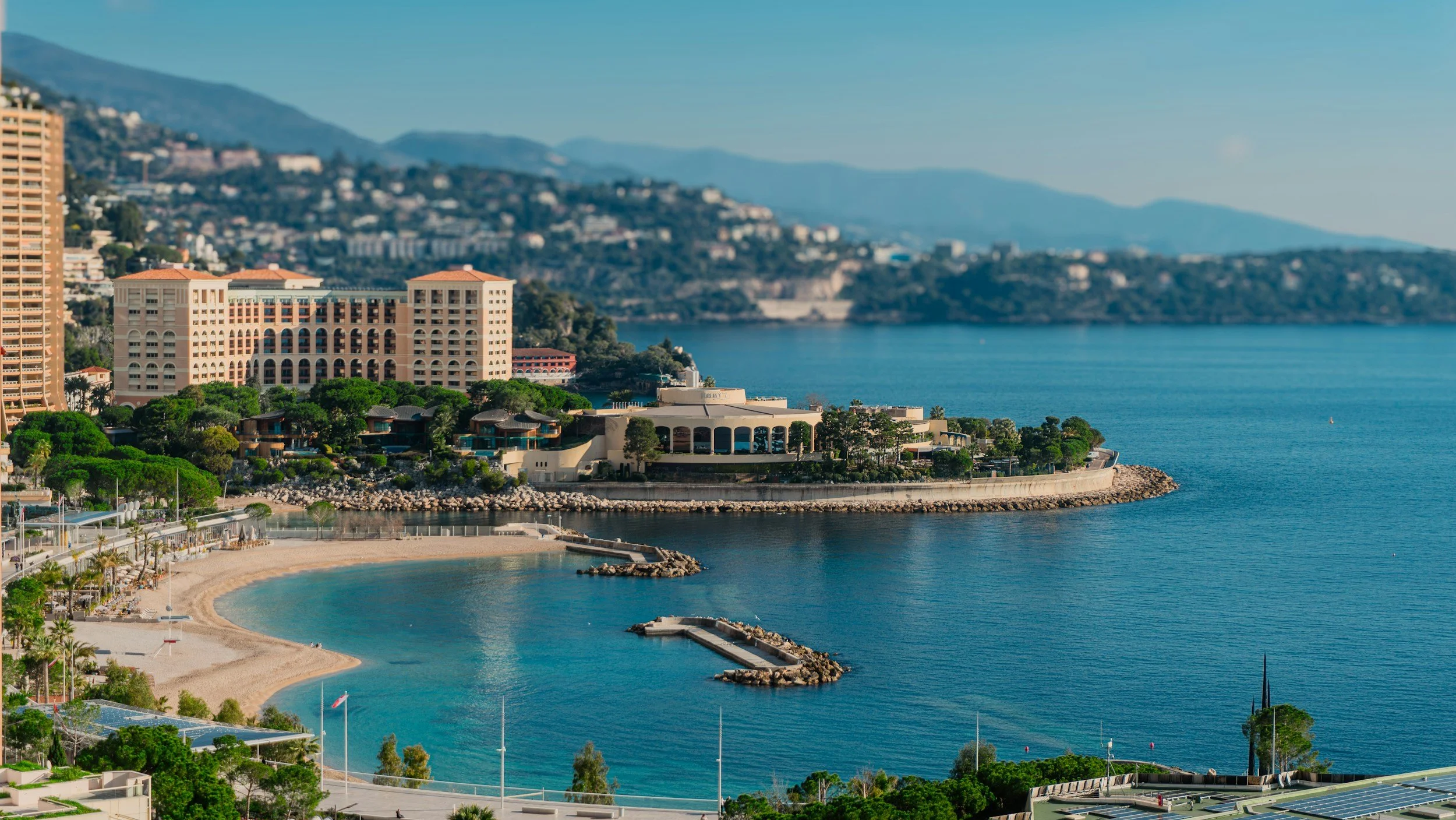 A coastal cityscape with modern buildings, a sandy beach, and calm blue water, with hills in the background.