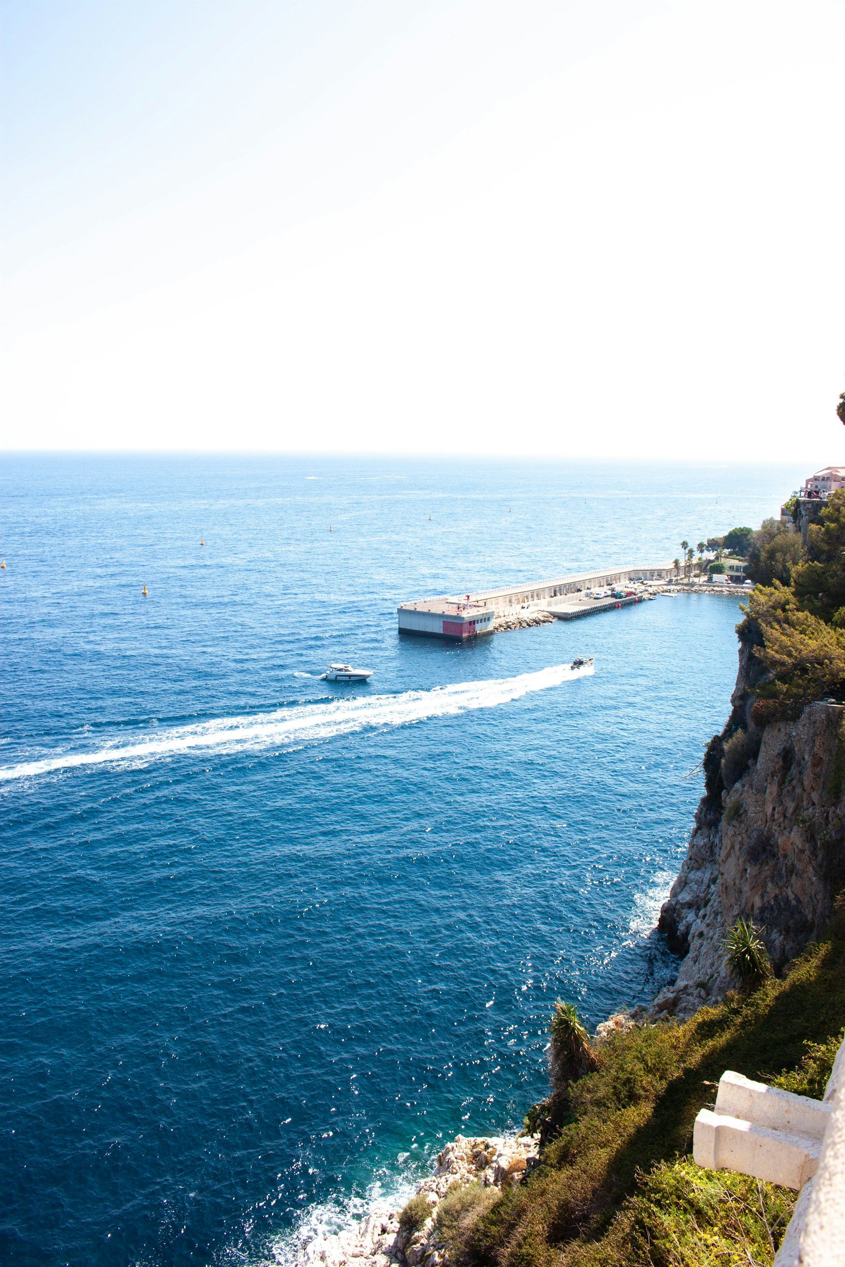 A view of the ocean from a coastal hillside with boats and a pier, and a white staircase on the foreground edge.