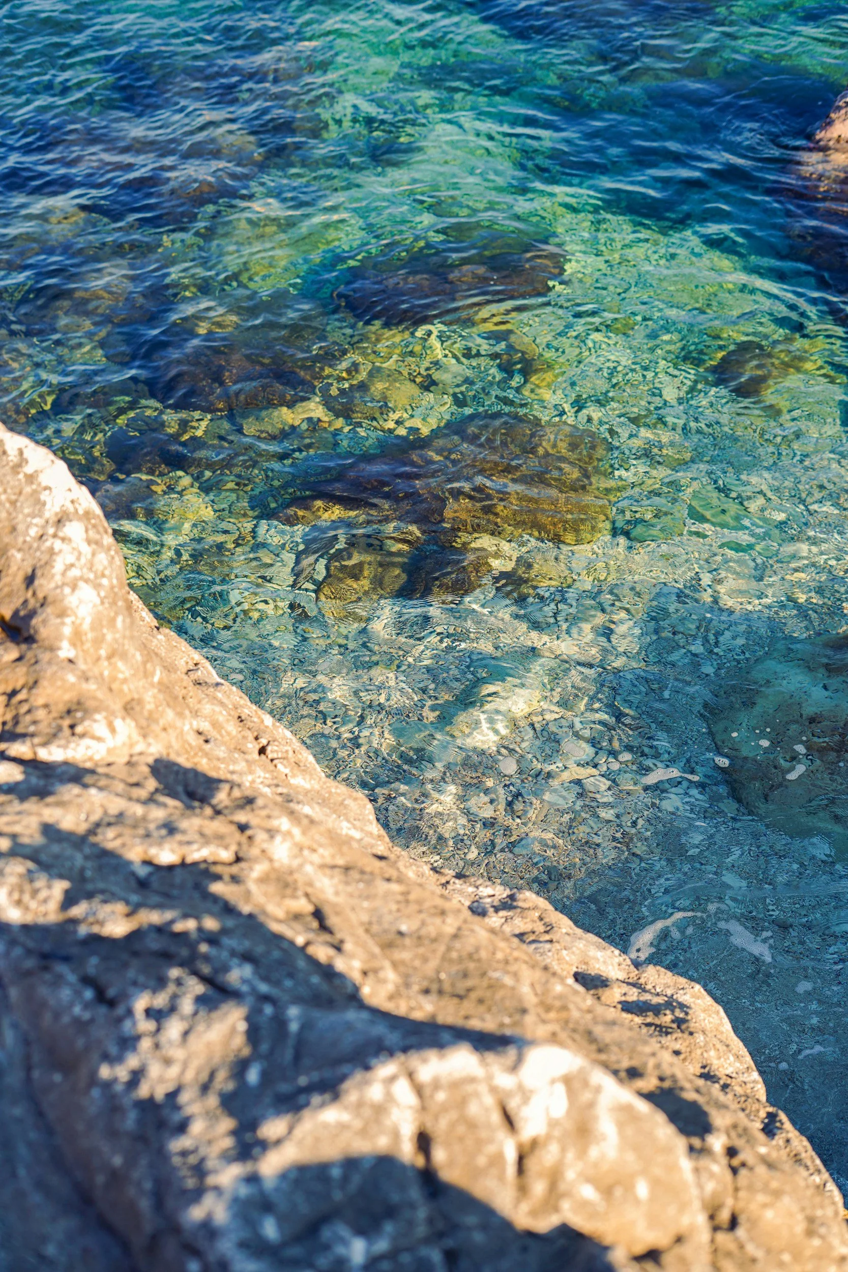 Clear blue water with rocks and pebbles visible below the surface, near a rocky shoreline.