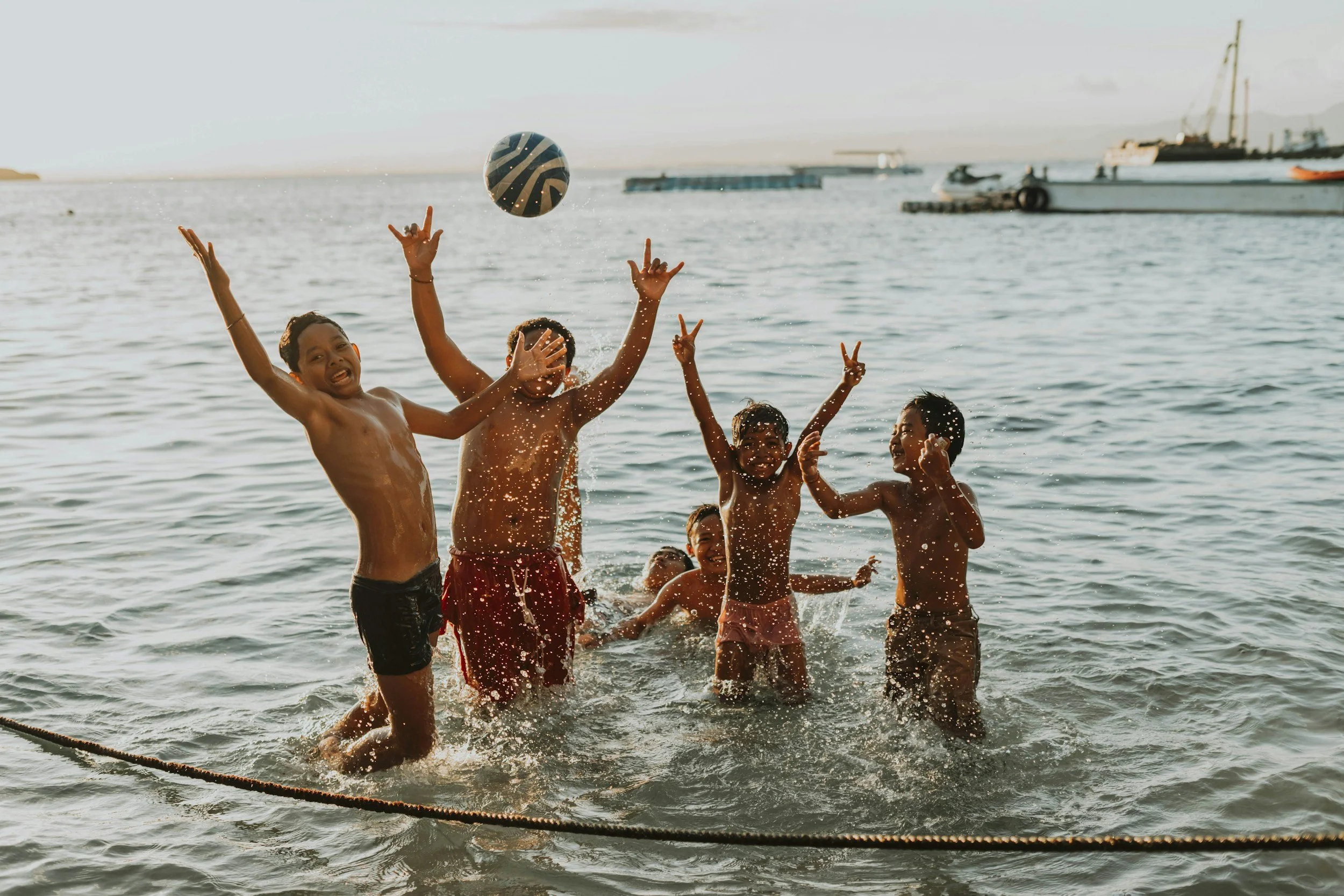Group of children playing and splashing water in a lake or ocean, with boats and a ship in the background during sunset.