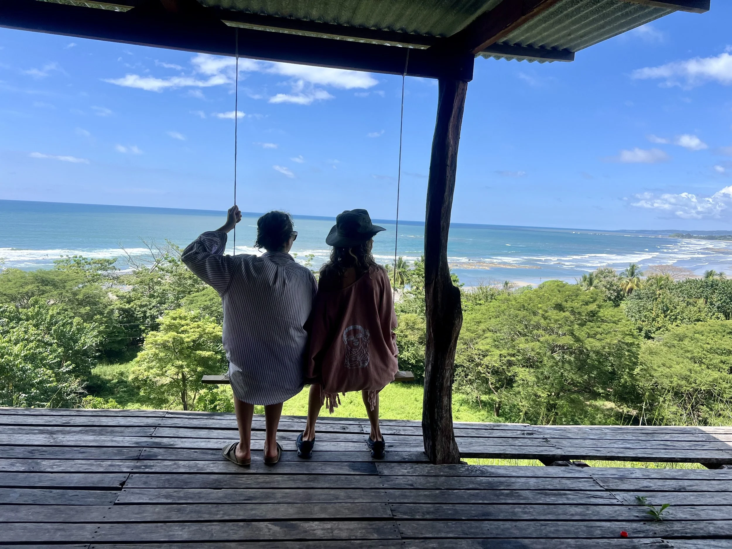 Two people sitting on a wooden deck overlooking a lush green forest and the ocean, with a blue sky and scattered clouds in the background.