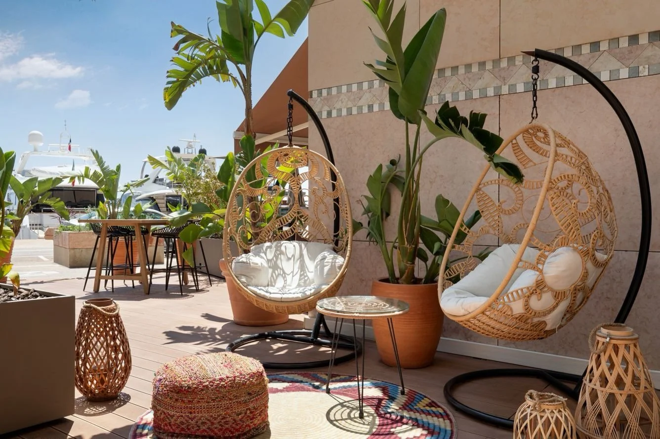 Outdoor patio with two hanging wicker chairs with cushions, a small round table, potted plants, and a view of boats docked at a marina under a blue sky.