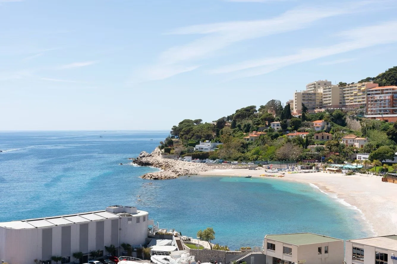 A coastal scene with a sandy beach, blue ocean, and hillside buildings including high-rise apartments and houses, under a partly cloudy sky.