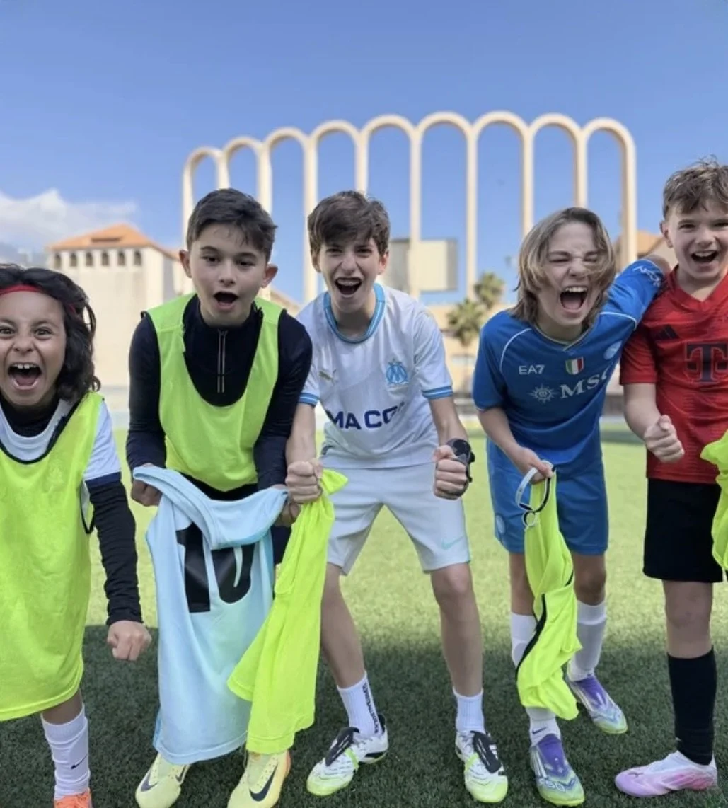 Group of children celebrating after a soccer game, smiling and holding their sports uniforms, with a large amusement park roller coaster in the background.