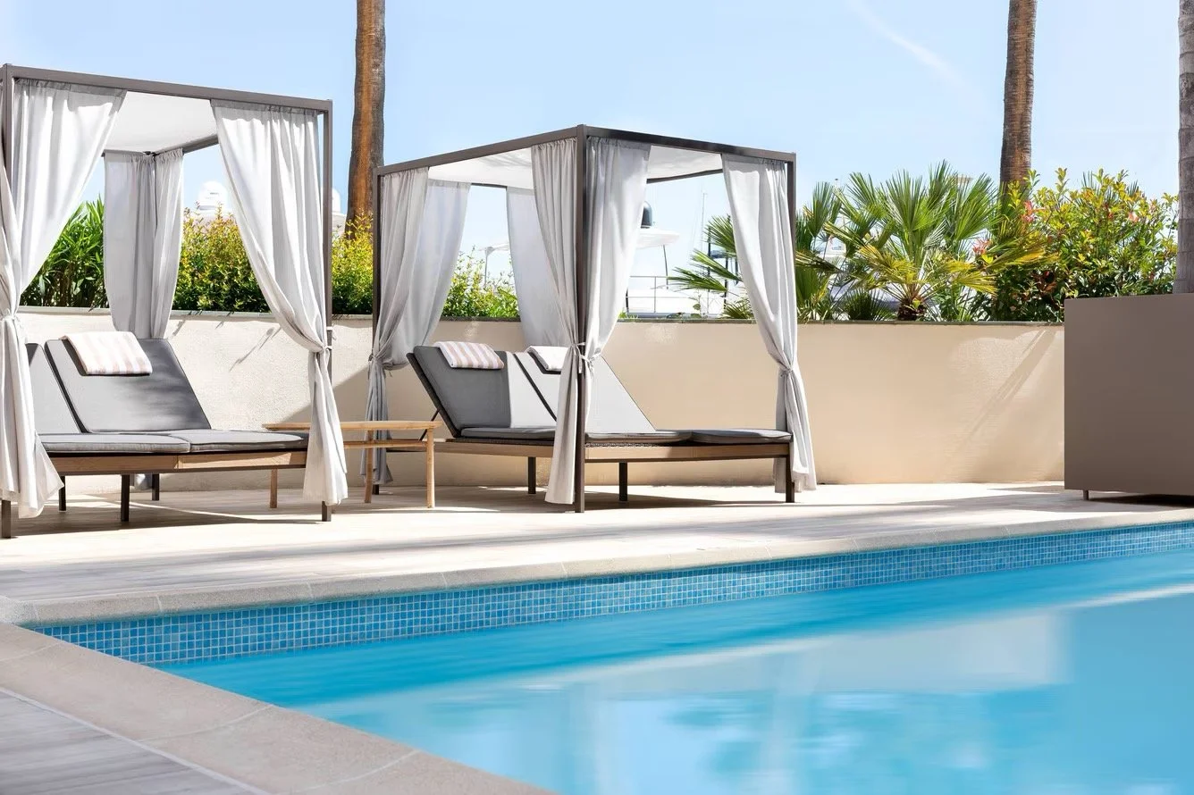 Poolside cabanas with white curtains and lounge chairs on a sunny day with palm trees and greenery in the background.