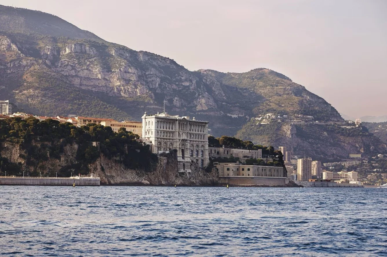 A coastal cityscape with a large, historic building on a rocky hillside overlooking the water, with mountains in the background and several high-rise buildings along the shoreline.