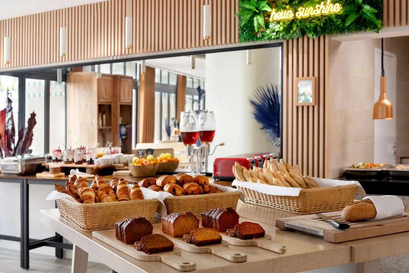 Breakfast buffet with croissants, sliced bread, cakes, and drinks in a modern dining area with wooden decor.