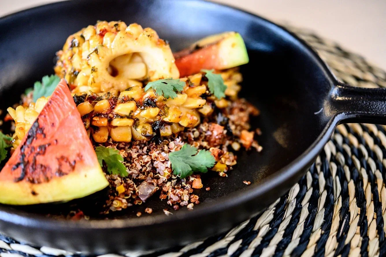 A black skillet containing grilled watermelon slices, quinoa, grilled corn, avocado slices, and garnished with cilantro leaves.