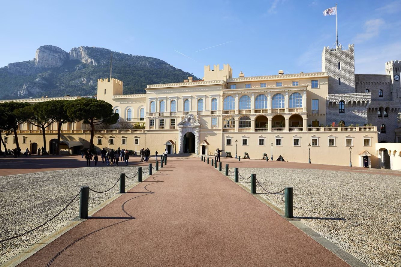 A large castle-like building with a castle wall and towers on the right side, situated against a mountain. The building has multiple floors with arched windows and a central ornate entrance. There are people gathered in front, some walking and some s