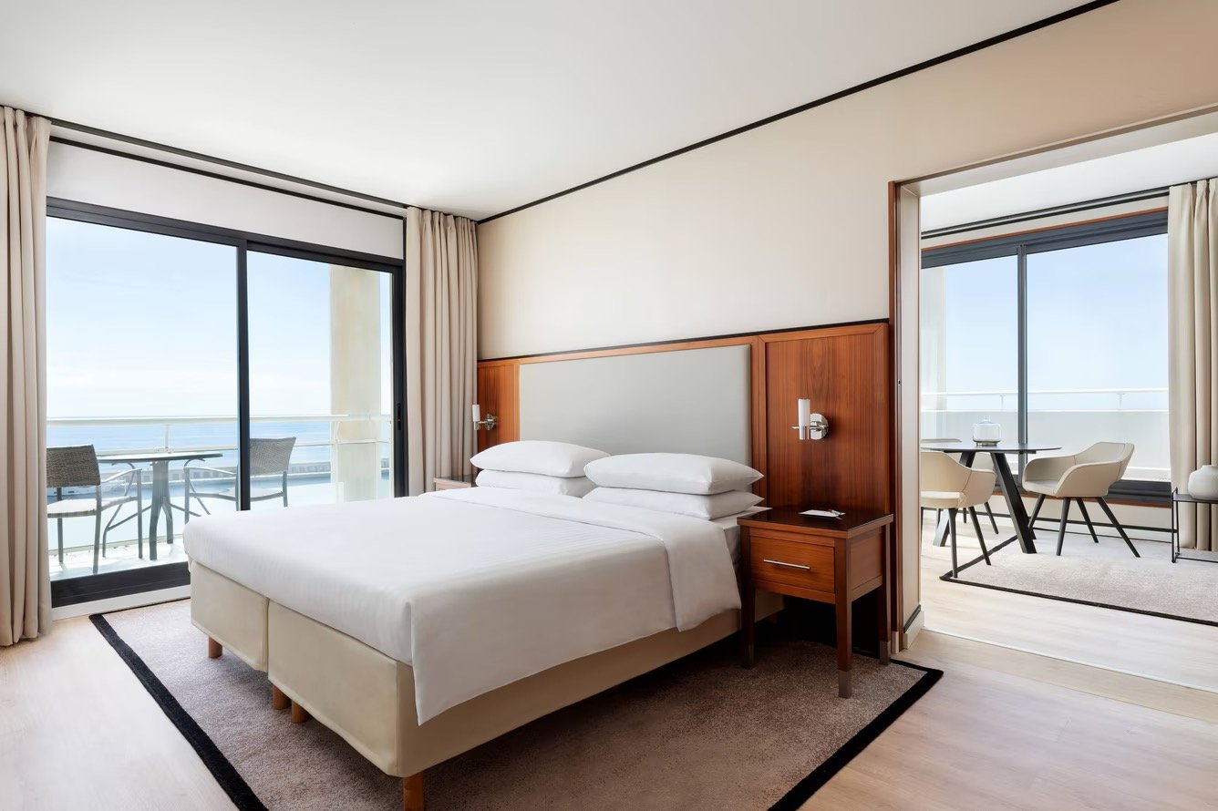 Hotel room with a large bed, sliding glass doors leading to a balcony with outdoor seating, and a separate sitting area with a table and chairs, overlooking the ocean.