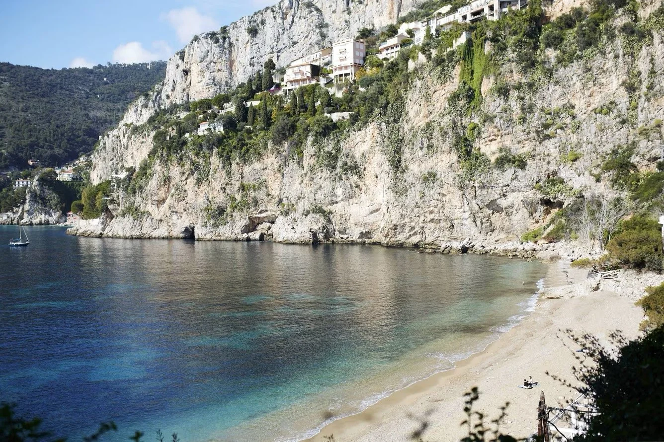 A peaceful beach scene with clear blue water, rocky cliffs, and houses perched on the hillside in the background.