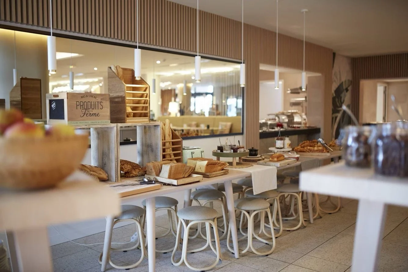 Breakfast buffet in a modern cafe with bread, pastries, jams, and utensils on white tables, seating chairs, and a background view of the cafe interior with warm lighting.
