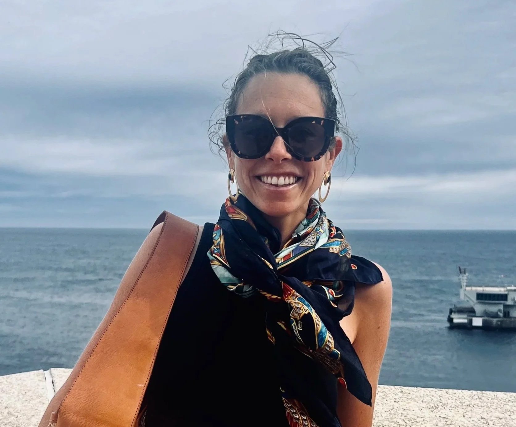 A smiling woman with big sunglasses, earrings, and a colorful scarf around her neck standing near the water with a pier and cloudy sky in the background.