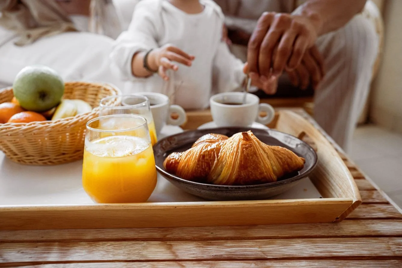 A breakfast tray with a croissant, a glass of orange juice, and cups of coffee, with people in the background having breakfast.