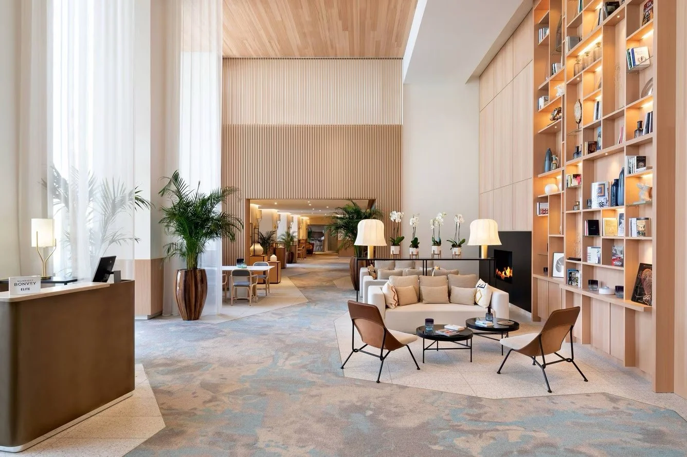 Modern hotel lobby with seating area, tall bookshelves, fireplace, large windows with curtains, potted plants, and contemporary decor.
