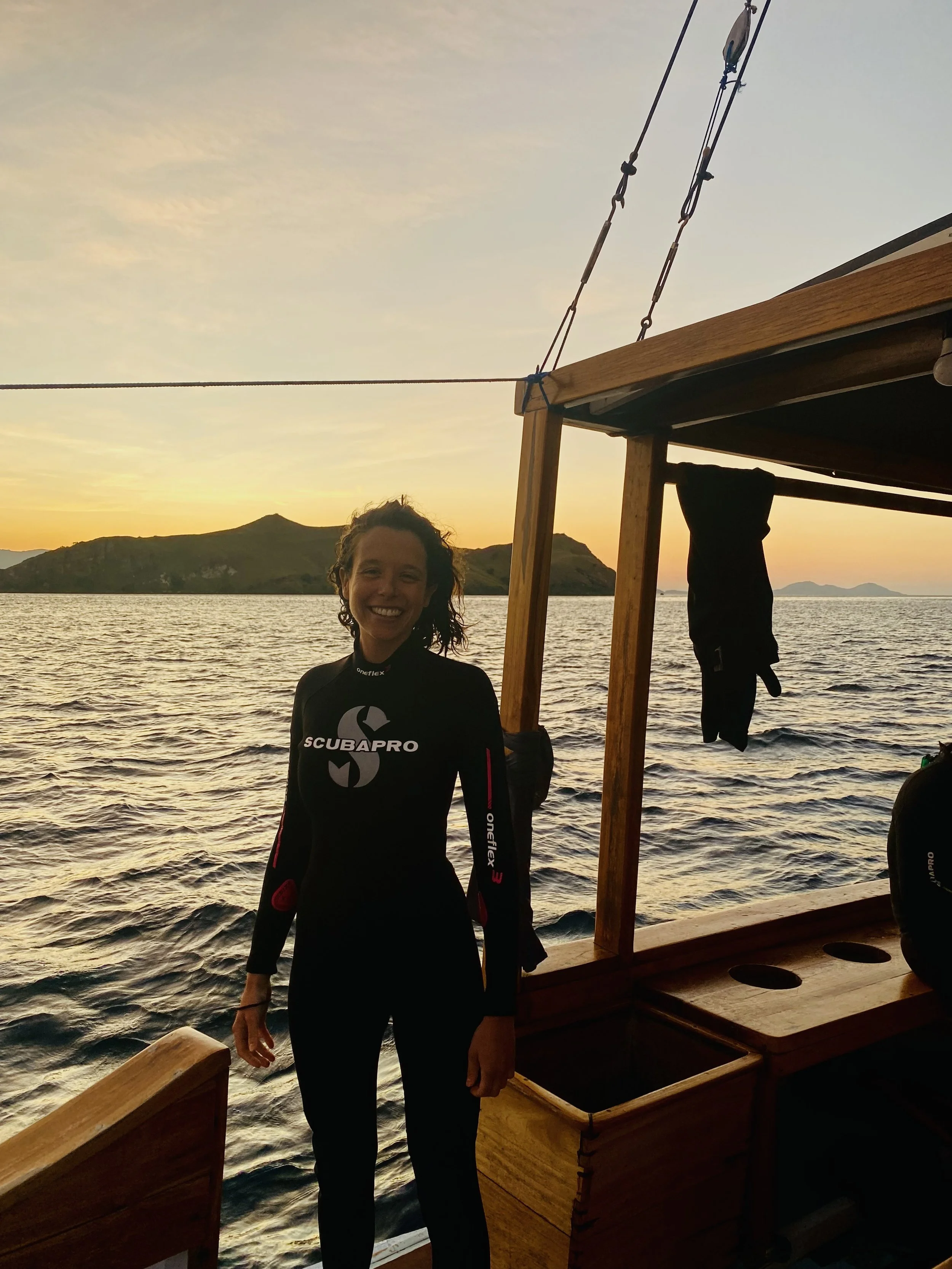 A woman smiling on a boat at sunset, with water and mountains in the background.