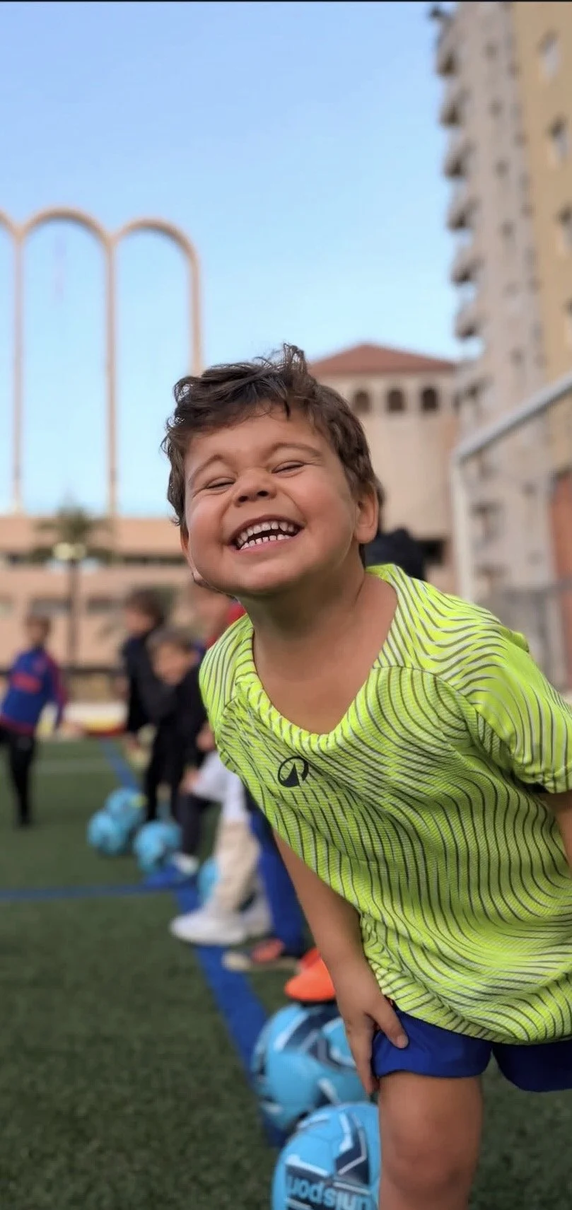 Smiling young boy in bright yellow sports shirt on a soccer field with kids and buildings in the background.