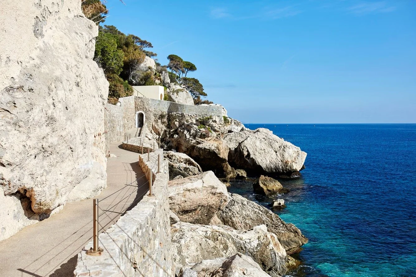 A coastal walkway along a rocky cliff with trees and white buildings, overlooking the blue ocean under a clear sky.