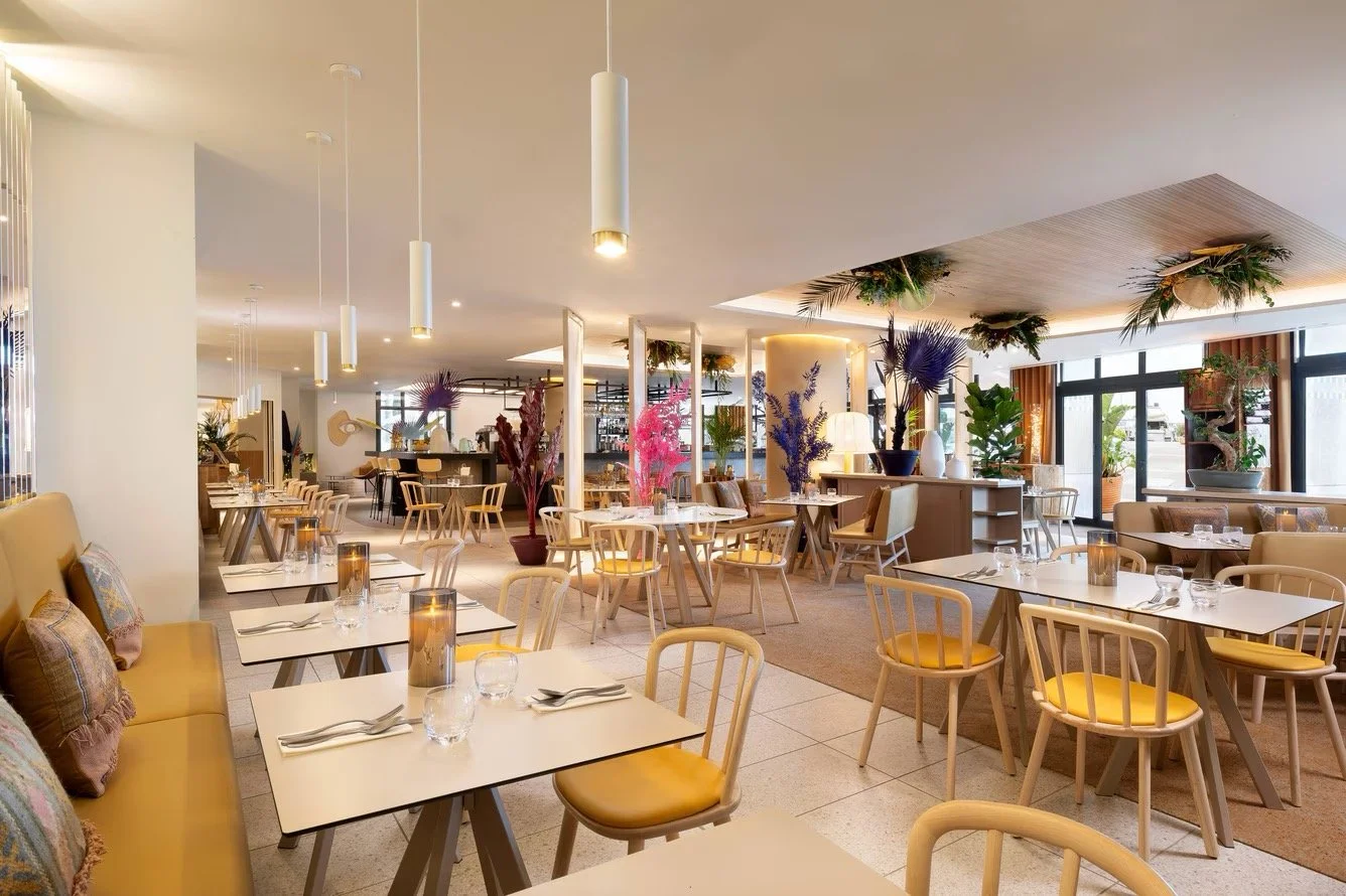 A modern restaurant with white tables, yellow cushioned chairs, and decorative plants and flowers in various vases, featuring floor-to-ceiling windows and warm lighting.