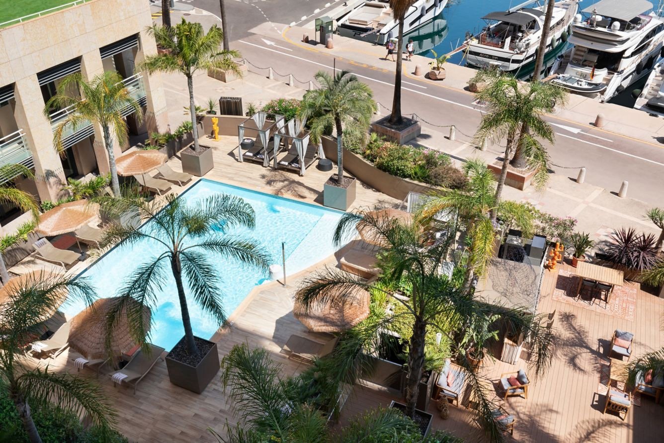 A swimming pool surrounded by palm trees and lounge chairs, with a view of a marina with boats docked along the waterway.