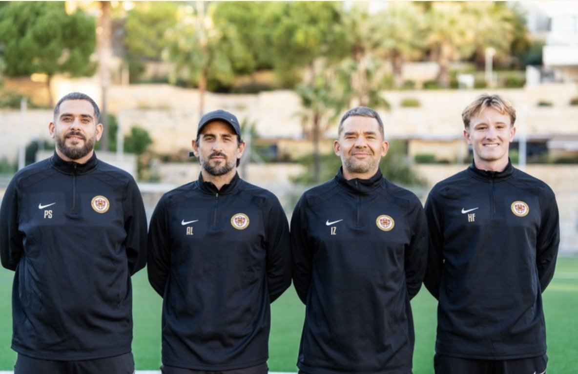 Four men wearing black sports jackets with a soccer team logo, standing on a field with trees and buildings in the background.