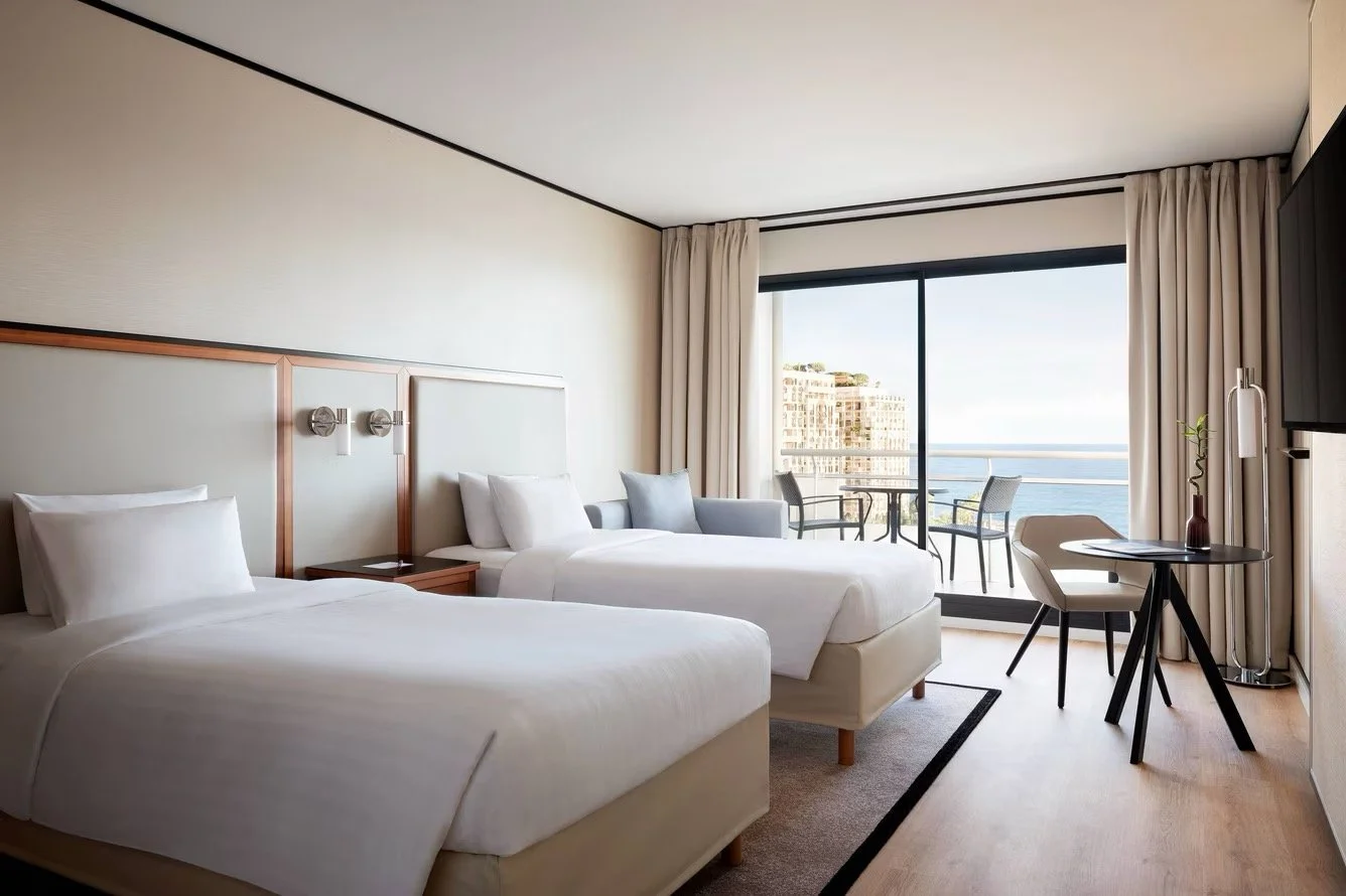 Hotel room with two beds, a balcony with outdoor seating, and ocean view.