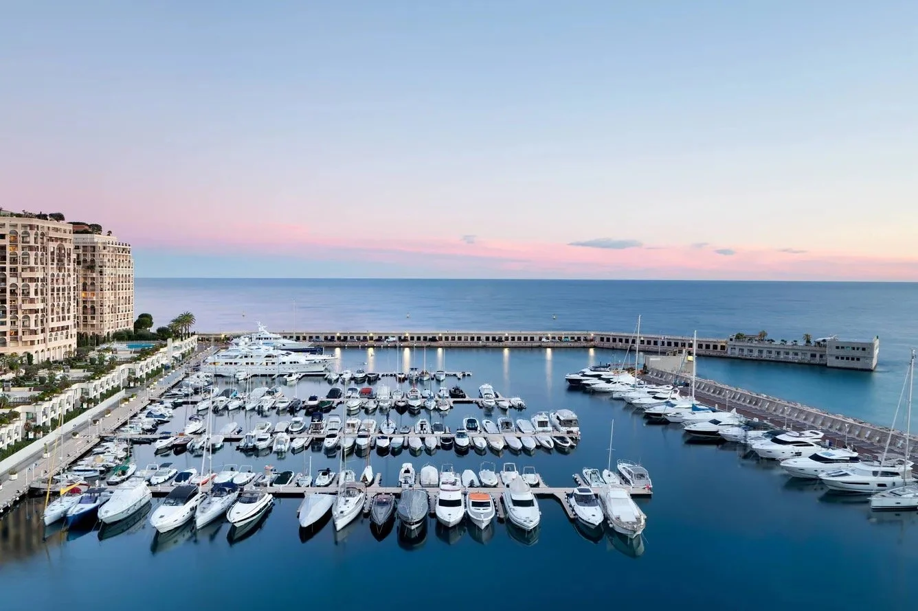 A marina with boats docked, surrounded by a breakwater, next to a residential building, with the ocean and pink sunset sky in the background.