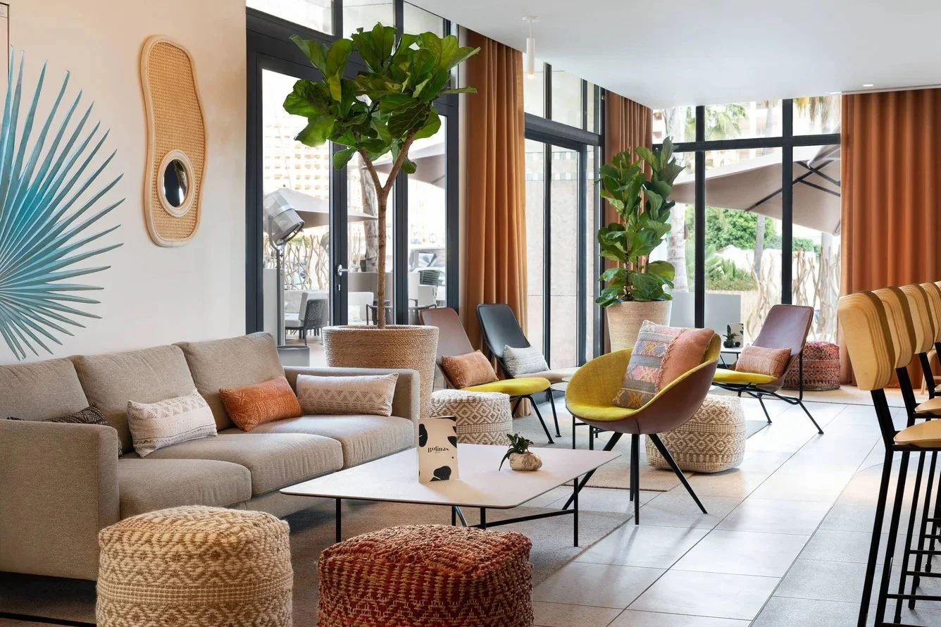 Modern indoor lounge area with a beige sofa, colorful accent chairs, large green potted plants, and floor-to-ceiling windows with orange curtains.