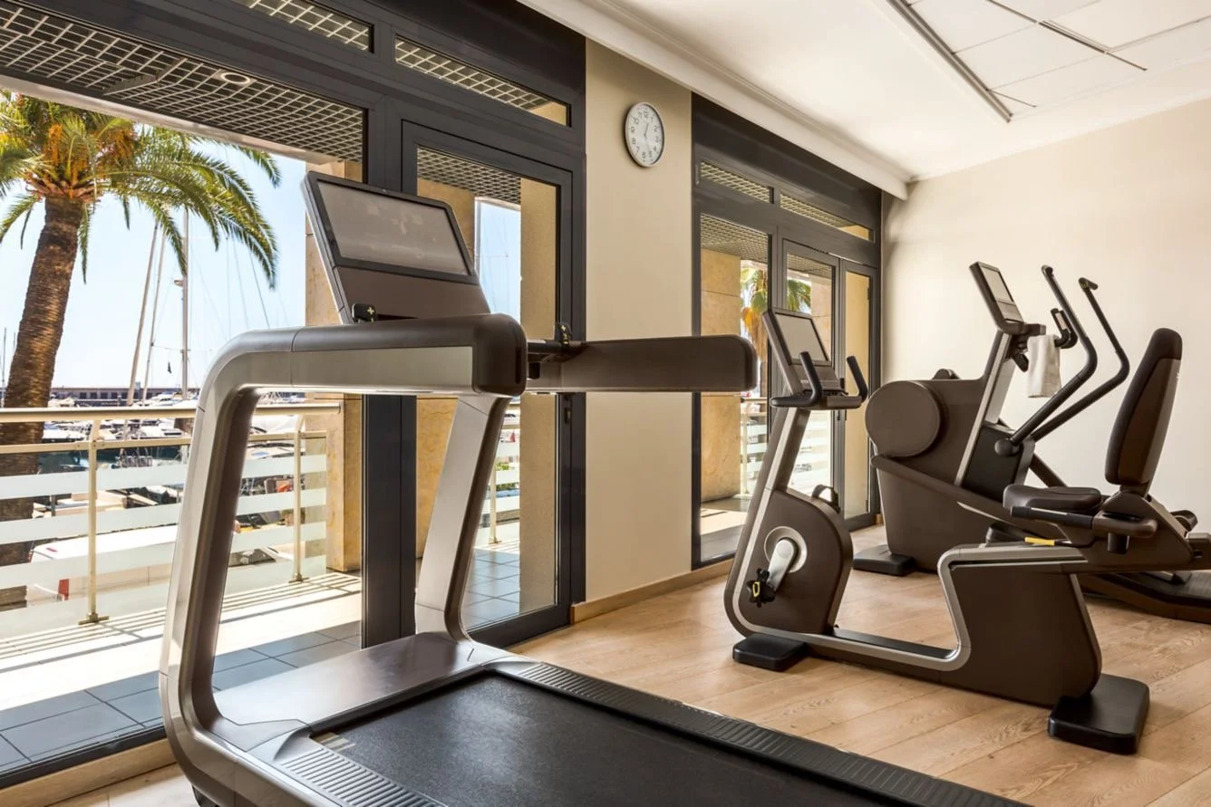 Indoor gym room with treadmills, an elliptical machine, and a stationary bike, large windows showing palm trees and a marina with boats outside.