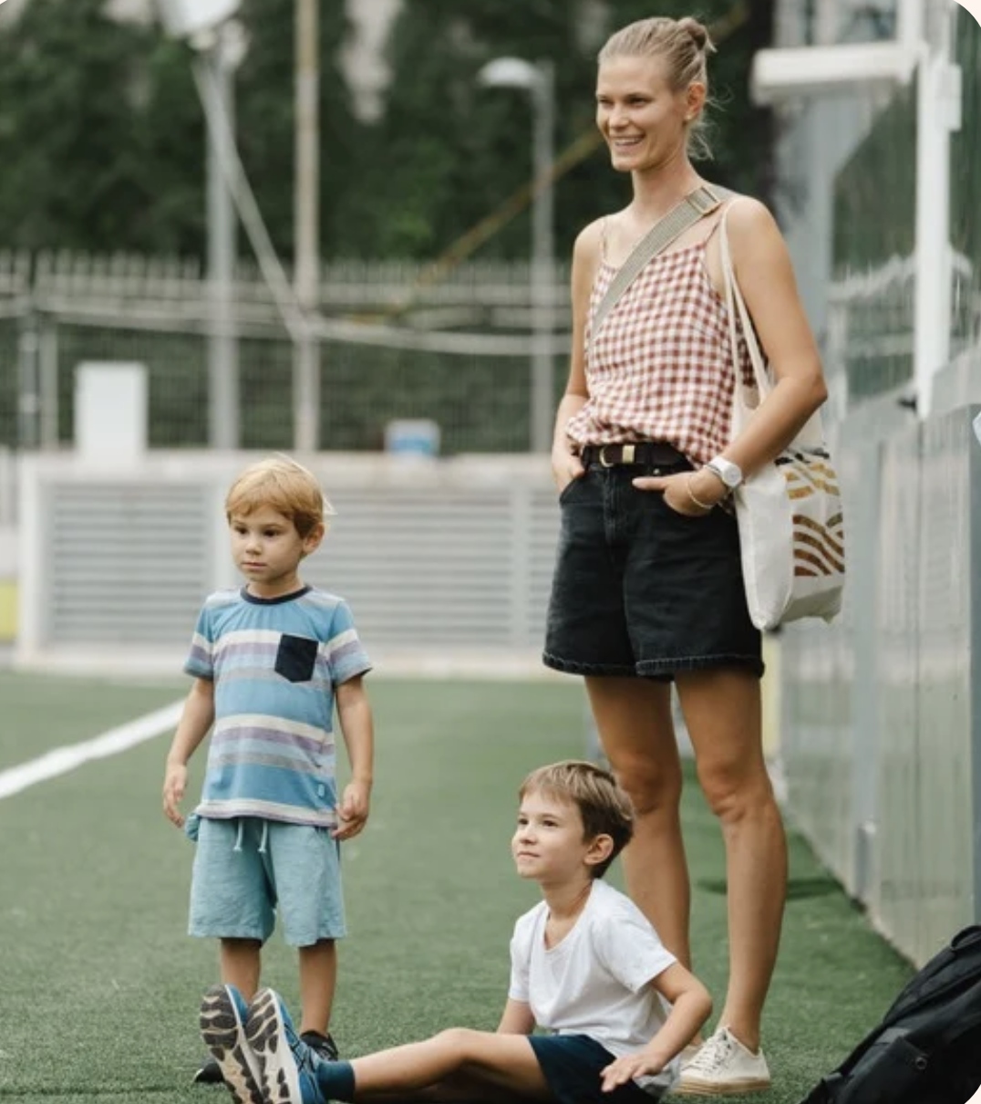 A woman standing on a tennis court with two young boys, one sitting on the ground and one standing, all casually dressed.