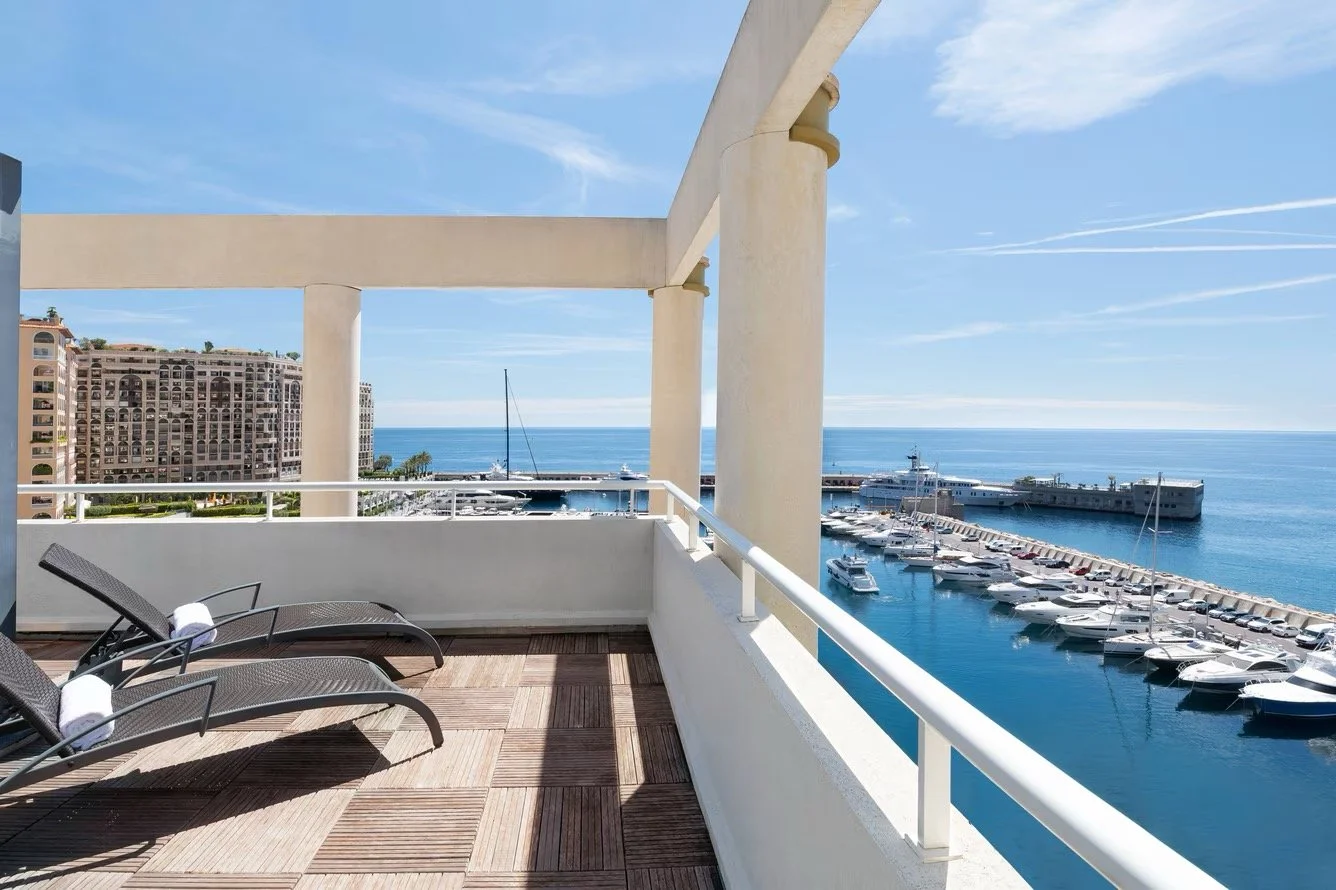 Balcony overlooking a marina with yachts, with apartment buildings and the ocean in the background, under a clear blue sky.