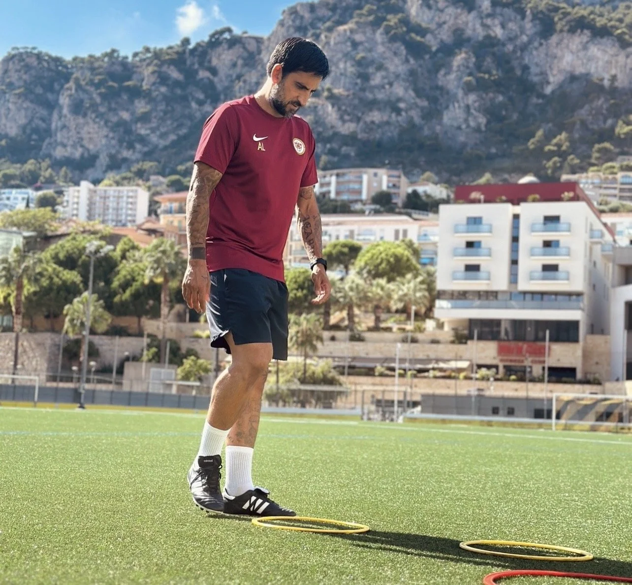 A man with tattoos in sportswear, including a maroon shirt and black shorts, practicing agility drills with yellow and red hoops on an outdoor field with city buildings and mountains in the background.