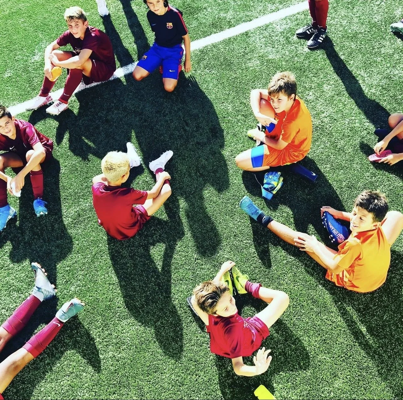 Children sitting on a soccer field in a circle, some wearing sports uniforms, sunlight casting long shadows.