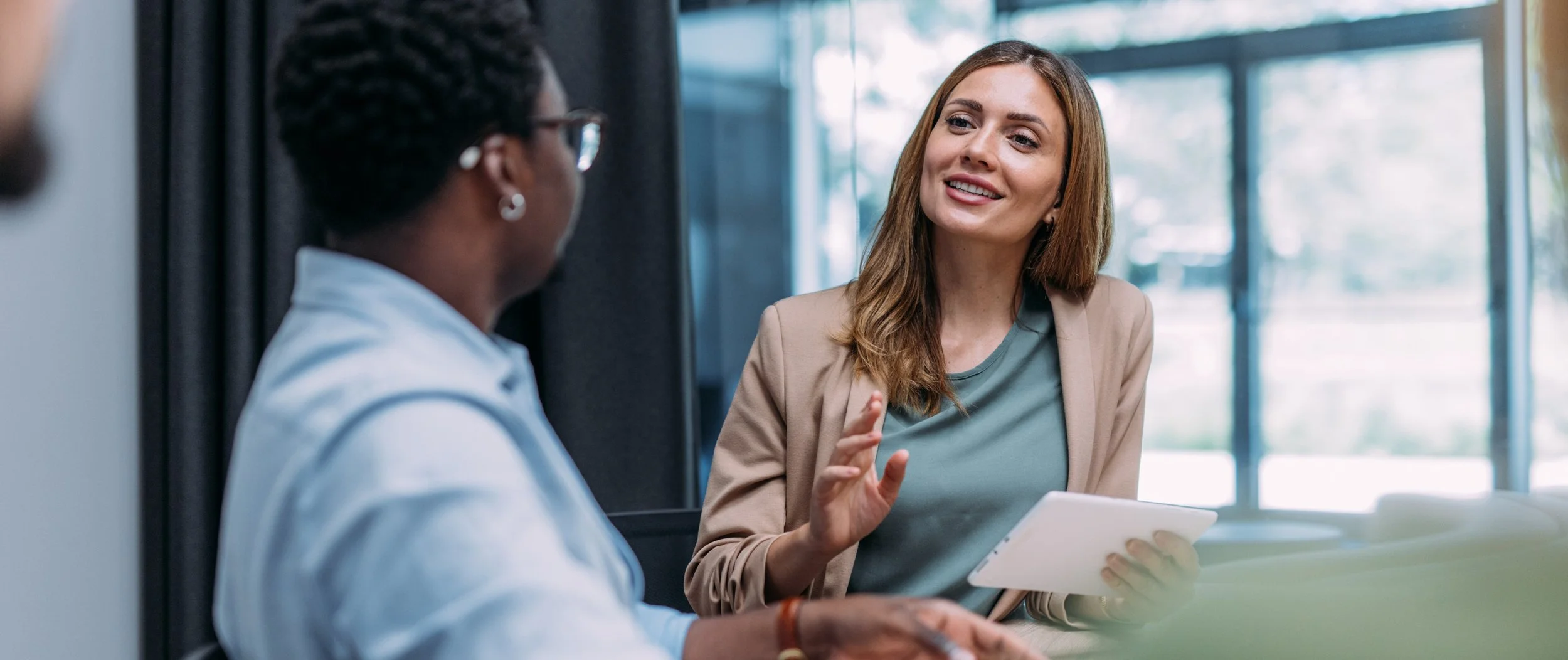 Two women sitting at a table engaged in conversation in a modern office space with large windows. The woman on the right is smiling, holding a tablet, and gesturing with her hand. The woman on the left has short hair, glasses, and is listening attentively.