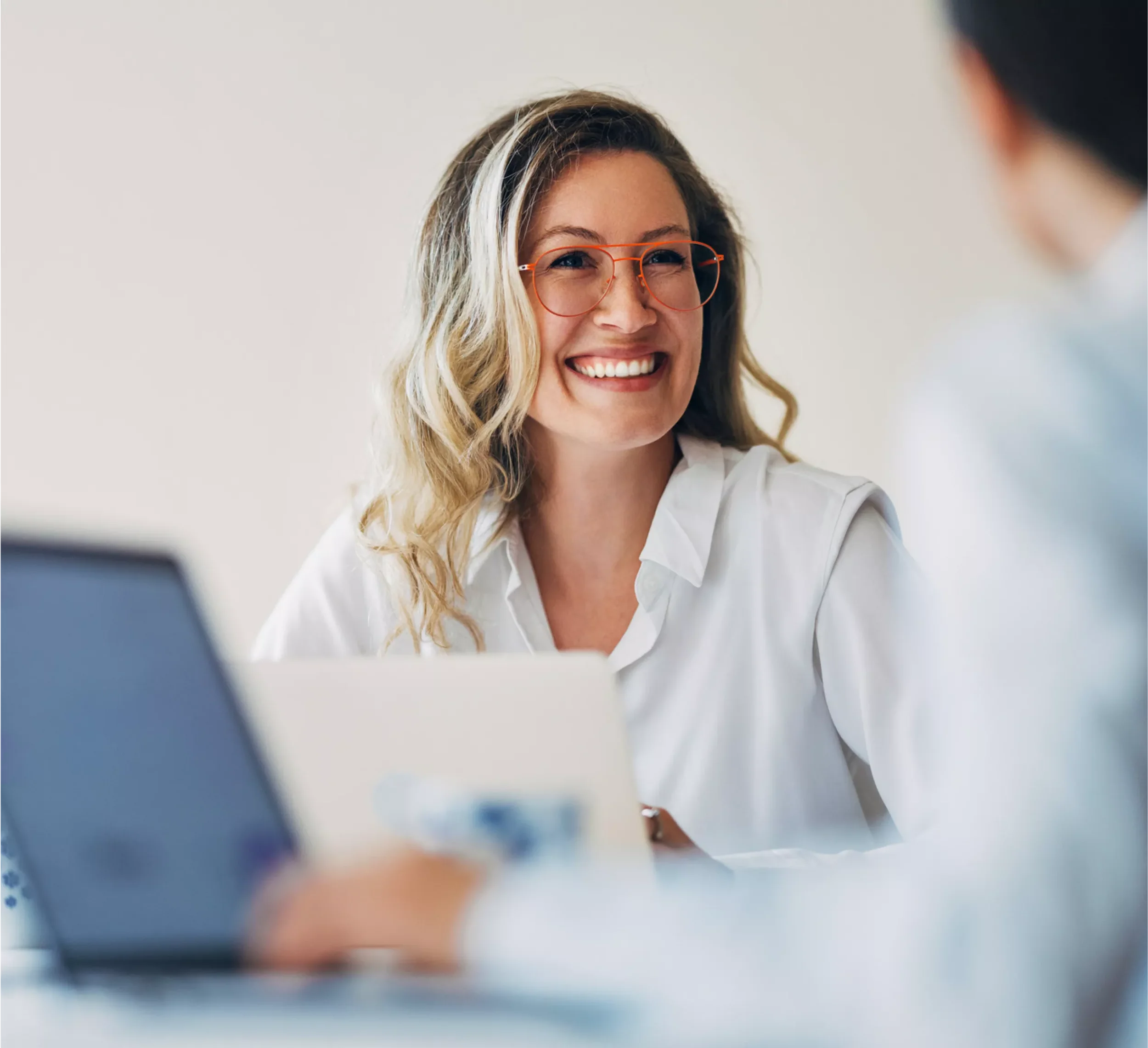 A woman with blonde curly hair, wearing glasses and a white shirt, smiling and engaging in conversation with another person at a table.