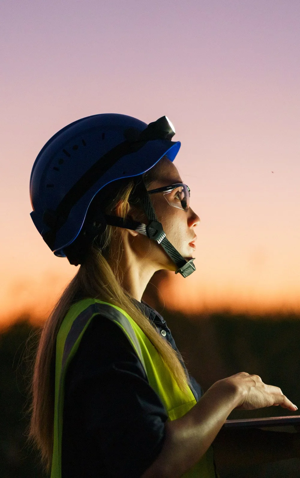 A woman wearing a blue safety helmet, glasses, and a yellow safety vest, looking into the distance during sunset or dusk.